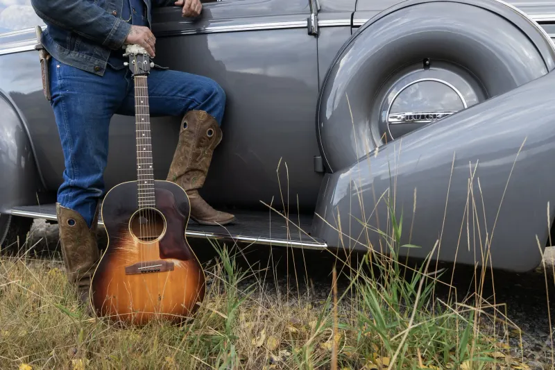 A guitar leaning on the running board of an old grey car, boots beside it.