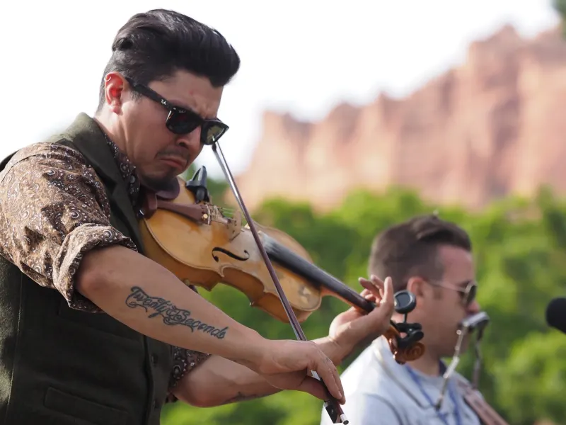 A fiddler in sunglasses playing outdoors, red cliffs behind.