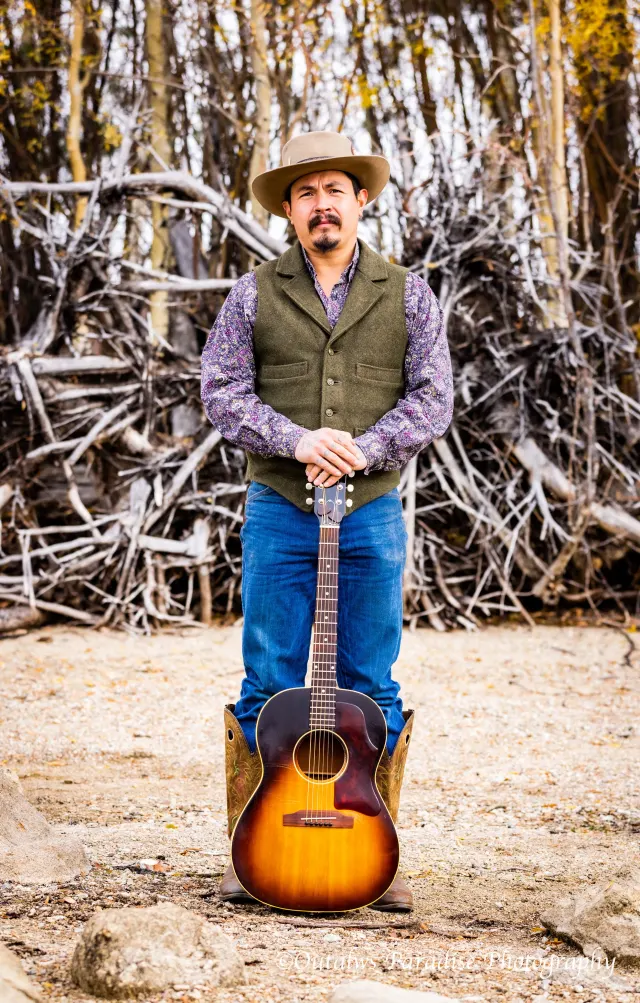 Rooster holding a sunburst acoustic guitar in front of a tall stack of weathered driftwood.
