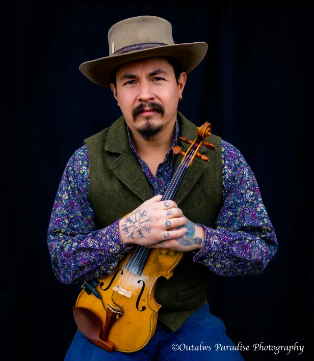 Reckless Rooster in a paisley shirt and vest, holding his fiddle against a black backdrop.
