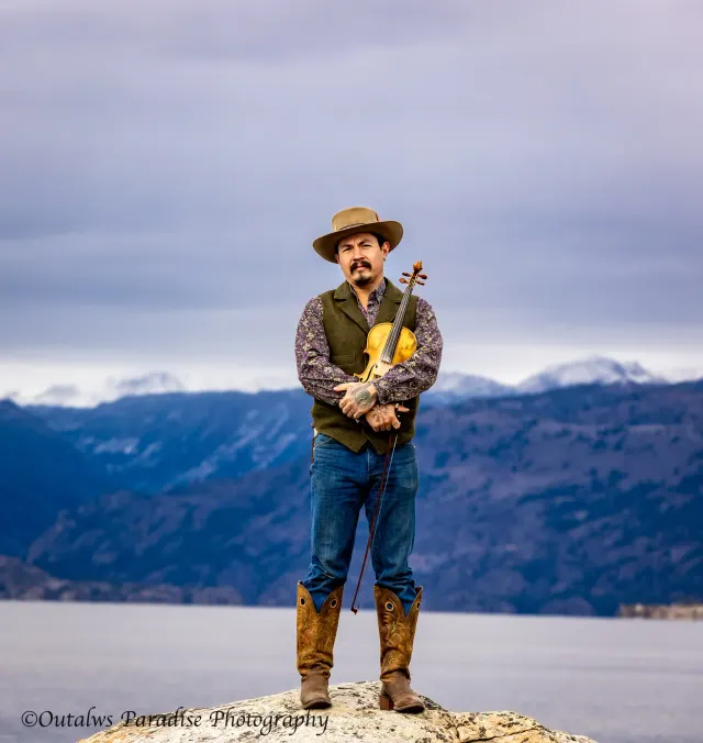 Rooster standing on a rocky shore with a lake and mountains behind him.